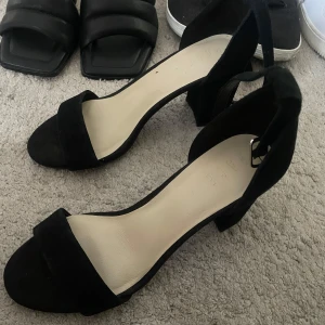 Svarta klackade sandaler med rem - Stilrena svarta sandaler från Dinsko med bred rem över tårna och justerbar rem runt vristen. Skorna har en chunky klack och är gjorda i mocka-liknande material. Perfekta för att lyfta vilken outfit som helst. Använda 1 gång