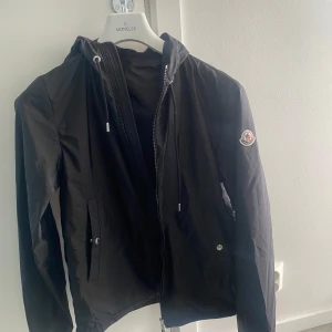 Svart vindjacka från Moncler - Svart vindjacka från Moncler med huva och snygga vita och röda detaljer på huvkanten. Jackan har dragkedja framtill, snörning i huvan och två fickor med knappar. Perfekt för dig som vill ha en stilren och sportig look. Äkta självklart! Storlek 1/S