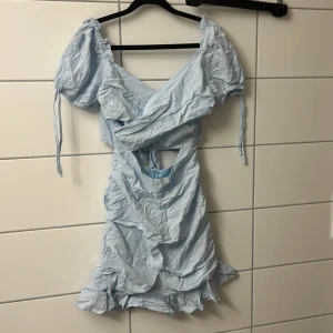 Ljusblå kort omlottklänning med volang - Supersöt ljusblå kort klänning med puffärmar, omlottdesign och volangkant nertill. Klänningen har knytning i ryggen och cut out-detalj i midjan. Perfekt för sommarens alla tillfällen och ger en fräsch vibe.( Skulle behöva strykas)