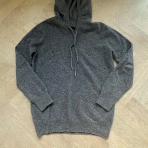 Grå hoodie i kashmir med snörning - Mysig grå hoodie i bland kashmir med huva och snörning. Tröjan har långärmad design och ribbade muddar vid ärmslut och nederkant. Perfekt för dig som gillar en enkel och stilren look med extra komfort. Tveka inte på att höra av dig!!!