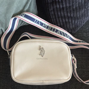 Vit axelväska från U.S. Polo Assn - Snygg vit axelväska från U.S. Polo Assn med silverfärgad logga framtill. Väskan har en bred, justerbar axelrem med blå och röda detaljer samt märkets namn tryckt längs hela remmen. Tillverkad i syntetmaterial och stängs med dragkedja.