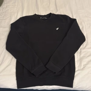 Svart sweatshirt från Pier One - En stilren svart sweatshirt från Pier One med rund hals och en liten vit broderad fågel på bröstet. Tröjan har långa ärmar och ribbade muddar vid ärmslut och nederkant. Perfekt för en clean och enkel look.