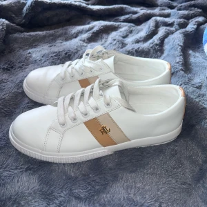 Vita sneakers från Ralph Lauren - Snygga vita sneakers från Ralph Lauren med beige detalj och RL-logga i guld på sidan. Skorna har rund tå, platt sula och klassisk snörning. Tillverkade i skinn för en clean och fräsch look, perfekta till jeans eller kjol. Storlek 38,5 Har använt de en gång. Priset kan diskuteras:)