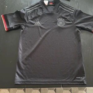 Svart Tyskland fotbollströja Adidas - Säljer en svart Tyskland fotbollströja från Adidas med silvriga detaljer och tre ränder på axlarna. Tröjan har korta ärmar med röd och gul kant, broderat landslagsmärke och Aeroready-material som andas. Perfekt för dig som älskar fotboll och vill ha en snygg matchtröja.