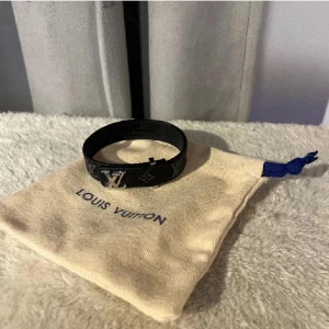  svart armband med monogramm mönstret ”LV” - Snyggt svart armband  med klassiskt monogrammönster och silverfärgad LV-detalj framtill. Tillverkat i skinn och levereras med originalpåse och ask. Perfekt accessoar för att lyfta din outfit med en lyxig touch.