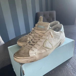 Philippe Model sneakers  - Snygga Philippe Model sneakers i beige mocka och skinn med ljusblå detalj på hälen. Klassisk låg modell med snörning och rund tå. Skorna har en ikonisk sköld på sidan och subtila kontrastsömmar. Perfekta för dig som gillar en avslappnad men trendig stil. Smutsiga så kan tvätta dem innan leverans.