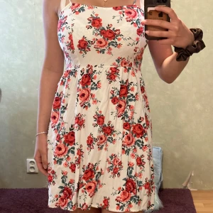 Vit blommig sommarklänning - Supersöt vit klänning med röda och rosa blommor, smala axelband och lätt utställd kjol. Perfekt för varma dagar med sitt luftiga tyg och romantiska mönster. Sitter fint i midjan och har en feminin siluett.