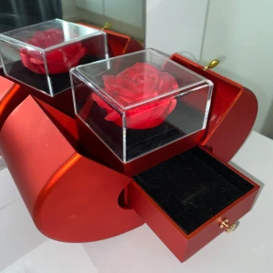 Röd hjärtformad smyckesask med ros - Unik smyckesask i metallicröd färg, formad som ett hjärta. På toppen finns en konstgjord röd ros i en transparent kub. Asken har en liten utdragbar låda med guldfärgad knopp, perfekt för att förvara ringar eller andra små smycken. Passar perfekt om ni vill använda det som present där ni lägger en typ av smycken i lådan.  Priset kan diskuteras!