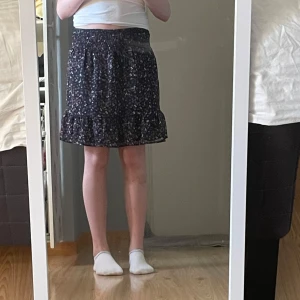 Blommig kortkjol  - Supersöt kortkjol med blommigt mönster i marinblått, rött och vitt. Kjolen har resår i midjan och en volang längst ner som ger en flowy känsla. Perfekt för dig som gillar en feminin och avslappnad stil.