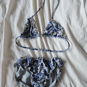 Blåvit volangbikini från Gina Tricot - Supersöt bikini från Gina Tricot i blått och vitt med volangdetaljer. Överdelen har trekantsmodell med smala axelband och spänne i ryggen. Underdelen har matchande volanger och mönster. Perfekt för strandhäng och poolparty! Jättefint skick, storlek 158/164