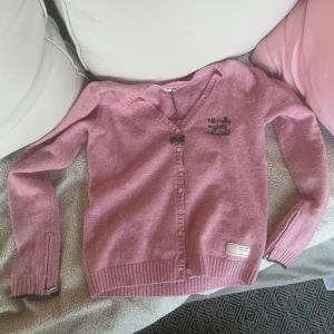 Rosa Odd Molly kofta med rosett - Supersöt rosa kofta från Odd Molly med v-ringning, knappar framtill och en liten svart rosett vid halsen. Broderad text på bröstet och en liten etikett nertill samt dragkedjor vid ärmarna. Köpt på Sellpy i bra skick men aldrig använd av mig💗