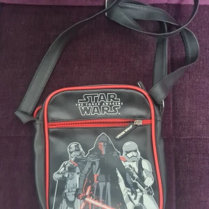 Star Wars axelväska med röd kant - Säljer en svart axelväska med motiv från Star Wars: The Force Awakens. Väskan har röd kant, justerbar axelrem och dragkedja framtill. Tryck med Kylo Ren och stormtroopers på framsidan. Perfekt för dig som älskar Star Wars och vill ha en unik accessoar.