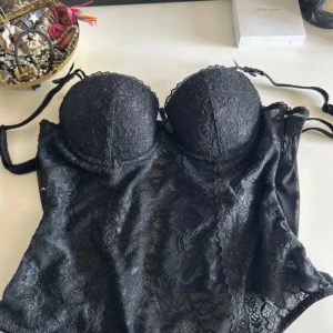 Svart spetskorsett från Etam - Supersnygg svart korsett från Etam med spetsdetaljer och vadderade kupor. Axelbanden är justerbara och tyget är genombrutet med vacker spets framtill. Perfekt för att lyfta din outfit med en edgy vibe.