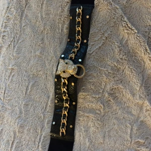 Svart bälte med guldkedja och leopardspänne - Unikt svart bälte i skinnimitation med ormmönstrad yta, dekorerat med guldfärgade nitar och en kraftig guldkedja. Spännet är en glittrig leopardhuvud med strass och ringdetalj. Perfekt för att lyfta vilken outfit som helst.