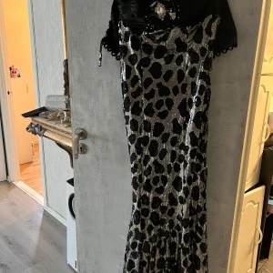 Lång glittrig leopardklänning - Snygg långklänning med glittrigt leopardmönster i svart och silver. Överdelen är svart med dekorativ brosch och korta ärmar. Klänningen har en figurnära passform och är perfekt för dig som vill sticka ut med en glamorös look.