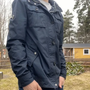  J.Lindeberg Jacka  - Snygg mörkblå vindjacka från J.Lindeberg med klassisk krage, flera fickor med knappar och dragkedja. Jackan har en stilren look och är perfekt för dig som gillar enkel men cool stil. Långärmad och tillverkad i polyester. Jackan har även en luva i kragen du kan ta ut eller ha kvar. 