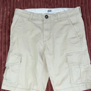 Beige cargoshorts från Jack & Jones - Snygga beige cargoshorts från Jack & Jones med klassiska cargofickor på sidorna och bälteshällor. Perfekta för sommaren med sin avslappnade passform och stilrena look. Gjorda i mjuk bomull som känns skön mot huden.