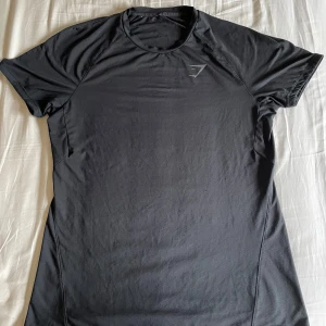 Svart Gymshark t-shirt i funktionsmaterial - Svart t-shirt från Gymshark med diskret logga på bröstet. Tillverkad i ett lätt och stretchigt funktionsmaterial som andas, perfekt för träning. Klassisk rund halsringning och korta ärmar för en sportig look.