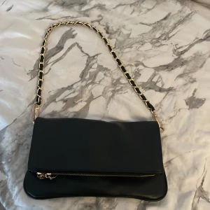 Svart axelväska med guldkedja - Snygg svart axelväska i skinnimitation med guldfärgad kedjerem och dragkedja framtill. Väskan har ett stilrent och modernt utseende, perfekt för att lyfta din outfit. Magnetlås och rymligt innerfack gör den både praktisk och trendig.