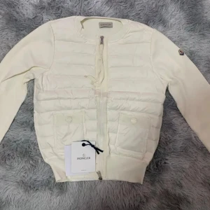 Vit cardigan jacka Moncler - Supersnygg vit cardigan jacka från Moncler med quiltad front och två fickor med knappar. Jackan har rund hals, dragkedja och Moncler-logga på ärmen. Materialmix med stickade ärmar och rygg samt vadderad framsida. Perfekt 