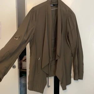 Olivgrön omlottjacka från Zara - Snygg olivgrön omlottjacka från Zara med långa ärmar och coola detaljer som snörning i midjan och metallring på ärmen. Jackan har en avslappnad passform och faller snyggt framtill. Perfekt för dig som gillar stilrena och trendiga plagg.