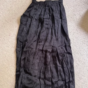 Svart långkjol med diskret mönster - En svart långkjol med subtilt mönster och lätt rynkad design. Kjolen har resår i midjan och ett flowy fall som ger en avslappnad men ändå stilren look. Perfekt för dig som gillar en klassisk vibe med en twist.