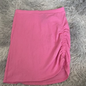 Rosa kortkjol med rynk från H&M - Snygg rosa kortkjol från H&M med rynkade detaljer på sidan och omlottkänsla. Kjolen är gjord i ett lätt och stretchigt polyestertyg som känns bekvämt och passar perfekt till en trendig look.