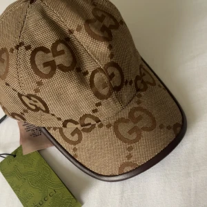 Gucci  keps beige (modell jumbo)  - Snygg beige keps från Gucci med klassiskt GG-mönster i brunt. Kepsen har justerbar rem i brunt skinn baktill och detaljer i guld. Tillverkad i Italien och har en exklusiv känsla med canvasmaterial och läderdetaljer.