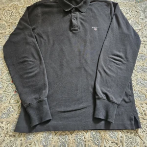 Svart långärmad pikétröja från Gant - Klassisk svart långärmad pikétröja från Gant med broderad logga på bröstet. Tröjan har krage, knappar framtill och ribbade muddar vid ärmsluten. Ett litet hål som man kan se på bilden, men som knappast syns eller märks. 