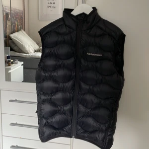Svart dunväst Peak Performance - Snygg svart dunväst från Peak Performance med quiltad design och hög krage. Västen har dragkedja framtill och diskret logga på bröstet. Perfekt att slänga över en hoodie eller tröja när det är kyligt ute.