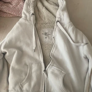 Vit teddyjacka med huva från H&M - Mysig vit teddyjacka från H&M med fluffigt foder, huva och dragsko. Jackan har dragkedja framtill och två stora fickor. Perfekt för kyliga dagar när du vill vara både varm och stilren.