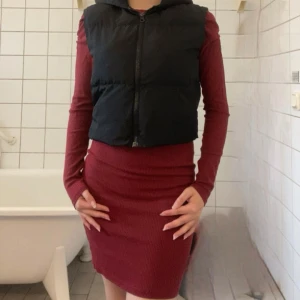 Svart croppad dunväst med huva NAKD - Säljer en svart, croppad dunväst med huva och dragkedja framtill. Västen är quiltad och har en enkel, stilren look som passar perfekt över en långärmad tröja eller hoodie. Perfekt för lager-på-lager-stil under kyliga dagar. Från NAKD