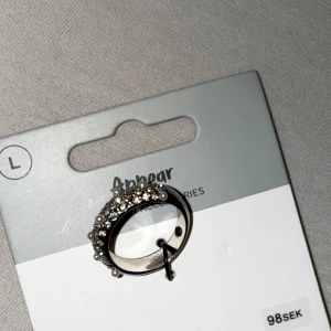 Silverring med pärlor och strass Appear - Bred silverfärgad ring från Appear, dekorerad med glittrande strass och små vita pärlor runt hela ringen. Perfekt statement-piece med lyxig känsla och snygg bling-effekt. Storlek L.