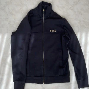 Svart tröja från BOSS - Svart tröja från BOSS med guldfärgad logga på bröstet och hel dragkedja framtill. Tröjan har hög krage, ribbade muddar och två fickor. Tillverkad i mjuk polyester, perfekt för en clean och sportig look. Tröjan har storlek S men passar M lika bra