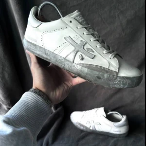 Vita sneakers från Premiata - Snygga vita sneakers från Premiata med grå detaljer och logga på sidan. Skorna har en unik grå sula med tryck och är tillverkade i skinn. Perfekta för dig som gillar stilrena och trendiga sneakers med en twist.