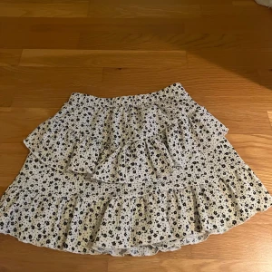 Vit blommig volangkjol - Supersöt kort kjol i vitt med små svarta blommor och flera volanger, inbyggda shorts.  Perfekt för dig som gillar en lekfull och trendig stil. Resår i midjan för skön passform. Hör av dig vid frågor, lägg prisförslag 💗