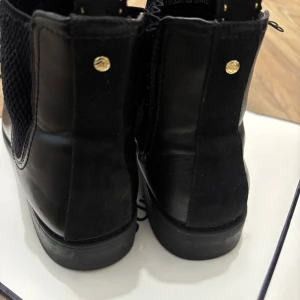 Svarta chelsea boots i skinn - Stilrena svarta chelsea boots i skinn med elastiska paneler på sidorna och diskreta guldfärgade nitar bak. Klassisk rund tå och låg klack, perfekt för en clean och tidlös look. Snygga detaljer och bekväm passform.