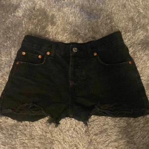 Svarta jeansshorts med slitningar - Snygga svarta jeansshorts med hög midja och råa, fransiga kanter. Shortsen har slitna detaljer framtill och klassiska fickor både fram och bak. Perfekta för en chill sommarlook.