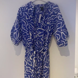 Blåvit mönstrad klänning Lindex S - Superfin blå klänning från Lindex i storlek S med vitt abstrakt mönster. Klänningen har korta puffärmar, v-ringning och knappar framtill samt ett matchande bälte i midjan. Perfekt för dig som gillar färg och vill sticka ut lite extra.