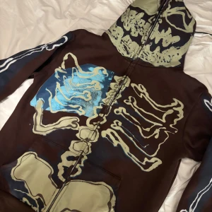 Brun four cactus hoodie med skelettmönster - Säljer en brun four cactus hoodie med hel dragkedja och huva. Tröjan har ett stort skelettmönster i beige och blått som täcker både fram- och baksidan samt huvan. Perfekt för dig som vill sticka ut med en unik och edgy stil. Använd en gång, storlek medium