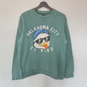 ASOS Oklahoma City sweatshirt green - Cool grön sweatshirt med tryck av en anka med solglasögon och texten 'Oklahoma City El Rino' på bröstet. Tröjan har rund hals, långa ärmar och ribbade muddar. Perfekt för dig som gillar streetstyle och vill sticka ut lite extra.