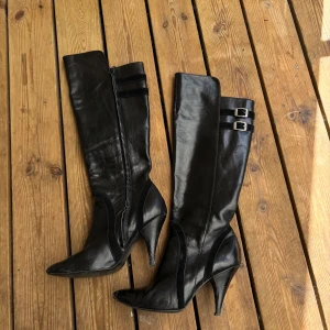 Svarta höga stövlar med klack - Snygga svarta stövlar i skinn med spetsig tå och hög klack. Stövlarna har dekorativa spännen upptill och dragkedja på insidan. Perfekta för dig som vill ha det extra till utekvällen eller bara vill spice up din outfit. 💋