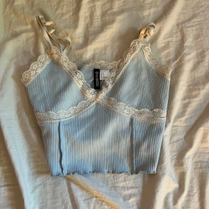 Ljusblå ribbad linnetopp med spets - Supersöt ljusblå ribbad linnetopp med smala axelband och vit spetskant vid halsringning och under bysten. Croppad modell med vågig nederkant, perfekt för sommaren. Säljer då den inte kommer till användning längre!