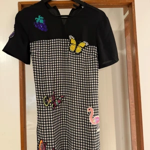 H&M Svart och vit rutig kortklänning med patches  - Kortklänning från H&M med svart överdel och svartvitt rutigt tyg nedtill. Klänningen har coola patches som fjäril, flamingo, vindruvor och texten 'Girl crew'. Knytband i midjan och korta ärmar. Detta är i storlek 34 och för mig som vanligtvis har storlek S så känns den fortfarande lite lös för mig. 
