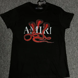 Svart Amiri t-shirt med röd orm - Svart t-shirt från Amiri med stor röd orm-grafik och vit logga på bröstet. Klassisk passform med rund hals och korta ärmar. Perfekt statement-plagg för dig som gillar edgy streetstyle.