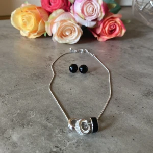 Set med halsband och örhängen i silver och svart - Snyggt smyckeset med ett tunt silverfärgat halsband med tre ringformade hängen, varav ett är glittrigt och ett är svart. Matchande svarta runda örhängen med stift. Perfekt för dig som gillar stilrena detaljer med lite bling.