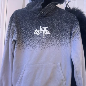 Svartgrå hoodie med SKATE-tryck - Snygg hoodie i svart och grått med coolt SKATE-tryck på bröstet. Tröjan har en unik färgövergång och knappdetalj vid halsen. Praktisk magficka och långärmad modell, perfekt för streetwear-stil.