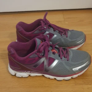 Grå och lila New Balance sneakers - Snygga New Balance 1080v5 sneakers i grått och lila med vita och röda detaljer. Ovandel i mesh och syntet för bra andningsförmåga, med klassisk N-logga på sidan. Skorna har snörning och en bekväm, stötdämpande sula som passar perfekt för träning eller långa promenader.