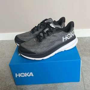 Svarta Hoka Clifton sneakers - Säljer ett par svarta Hoka Clifton sneakers med vit sula och svarta snören. Ovandelen är i mesh för bra andningsförmåga och komfort. Skorna har en dämpad sula som ger grym support vid löpning eller promenader.  Obs junior storlek, skulle säga att dom är mer som storlek 36. Skickas i original kartong 