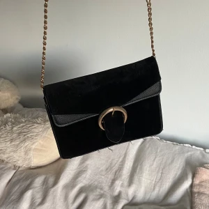 Svart mocka axelväska med guldkedja - Snygg svart axelväska i mocka med guldkedja som axelrem och ett stort guldfärgat spänne framtill